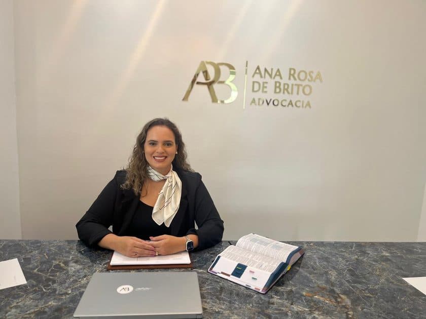 Ana Rosa de Brito sentada à mesa do escritório, com o letreiro Ana Rosa de Brito Advocacia ao fundo.